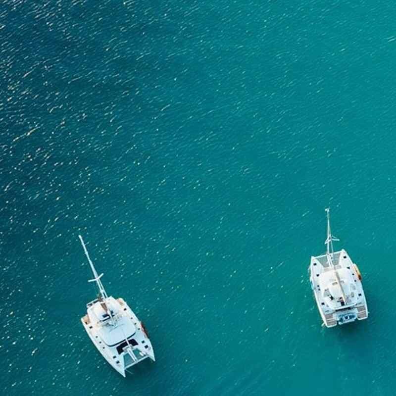 Vista aérea de dois veleiros ancorados sobre águas costeiras turquesa.